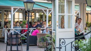 An outdoor seating area with wicker chairs and tables, surrounded by potted plants and a metal lamp post.