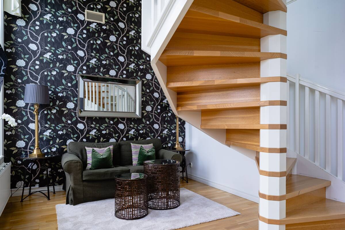 A staircase with wooden steps and white railings, a black floral wallpaper, a green sofa with cushions, and a white rug.
