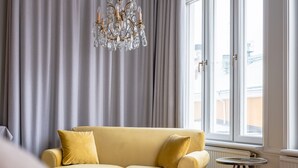 A hotel room with a bed, a yellow sofa, a chandelier, a window with curtains, and a radiator.