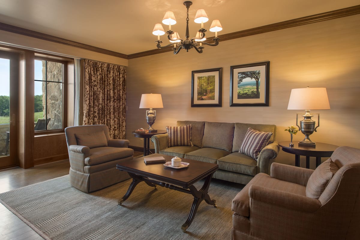A living room with a sofa, armchairs, a coffee table, and a chandelier. There are two lamps on side tables, and a window with curtains.