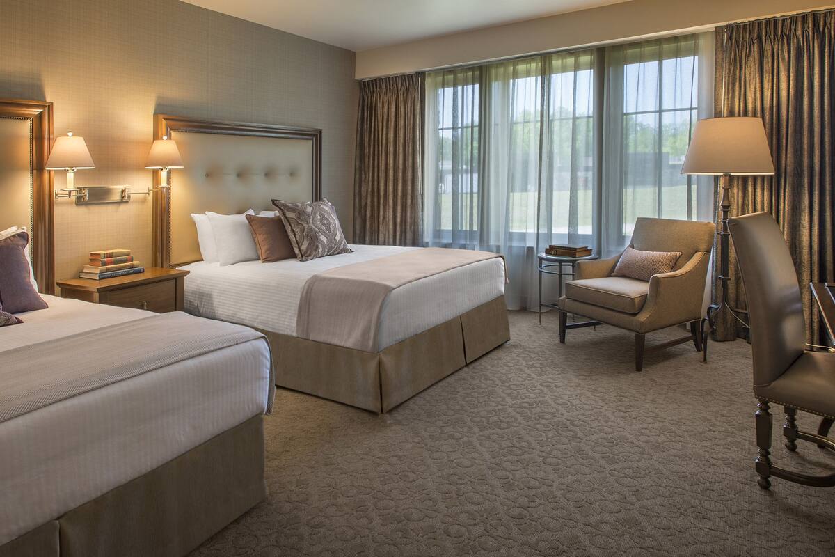 A hotel room with two beds, a chair, a desk, and a lamp.