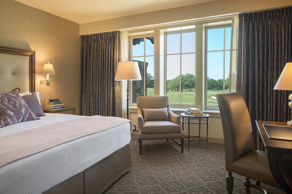 A hotel room with a large bed, two armchairs, a small table, and a view of a golf course through the windows.
