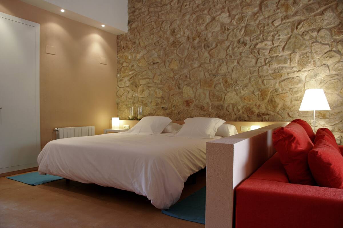 A bedroom with a stone wall, a bed with white bedding, a red sofa, and a nightstand with a lamp.