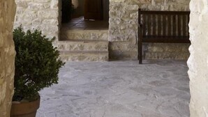 Pátio com paredes de pedra, um banco de madeira, uma planta em vaso e uma porta que dá para o interior.