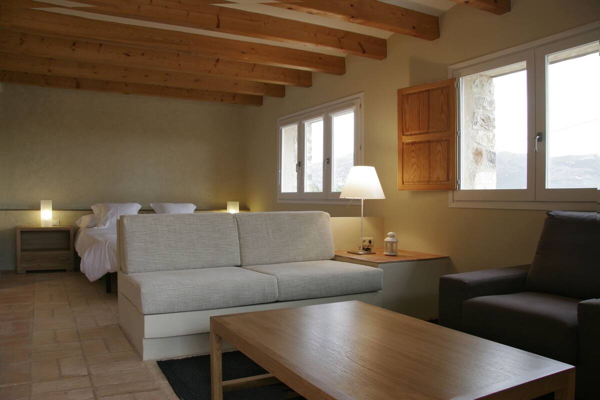 A cozy room with a sofa, a wooden table, a lamp, and a window with a view.