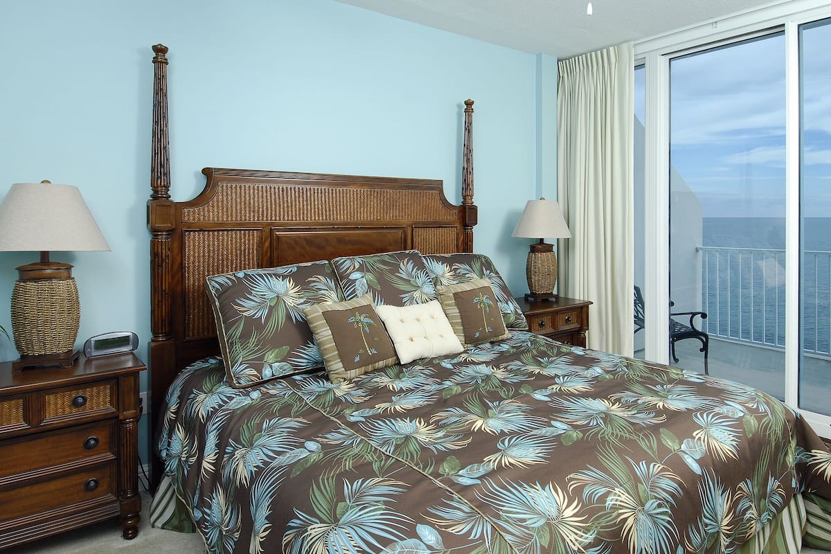 A bedroom with a four-poster bed, two bedside tables, a nightstand, a window with a view of the sea, and a balcony with a chair.