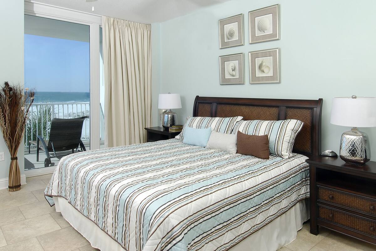A bedroom with a large bed, a nightstand, a lamp, and a view of the ocean.