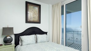 Un dormitorio con cama, mesita de noche, lámpara, cuadro enmarco y balcón con vista a la playa.