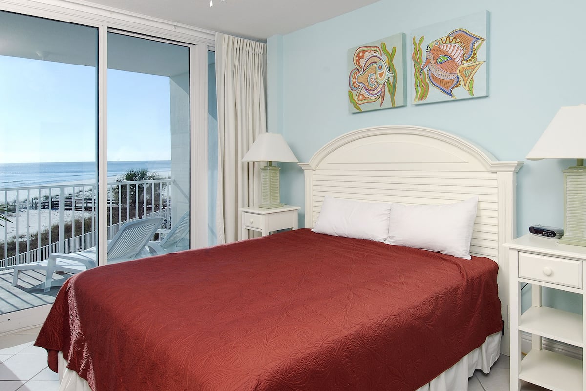A bedroom with a large bed, white nightstands, a balcony with a view of the beach, and two wall art pieces.