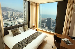 A hotel room with a large bed, a wooden desk, and a view of a cityscape and ocean.