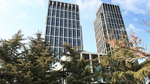 Twee moderne hoge gebouwen met een heldere lucht en weelderige altijdgroene bomen op de voorgrond.