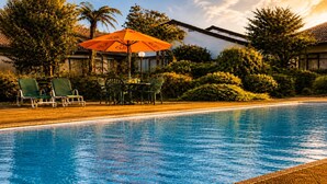 A swimming pool with lounge chairs and an umbrella, surrounded by greenery and a building. - Alpin Motel & Conference Centre (Rotorua)
