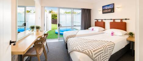 A hotel room with two beds, a desk, chairs, a small balcony, and a view of the outdoors.