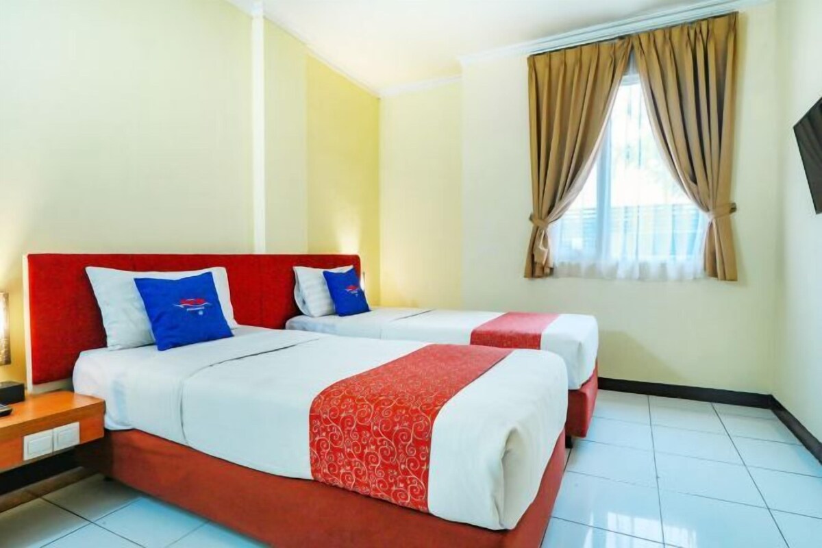 Quarto de hotel com duas camas, cada uma com roupa de cama vermelha e branca, cabeceira vermelha e janela com cortinas bege.
