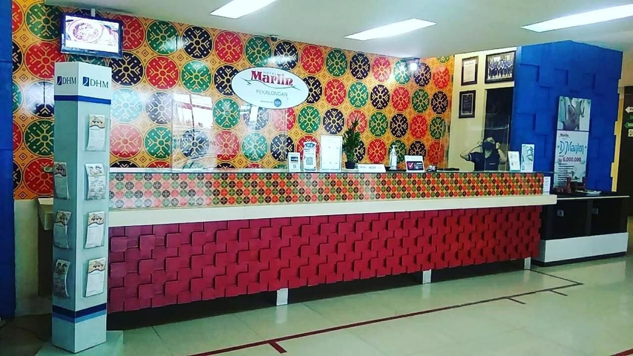 Un mostrador de recepción con una pared decorada con un patrón colorido, un cartel que dice "Marlin Pekalongan" y una pared azul con un cartel a la derecha.