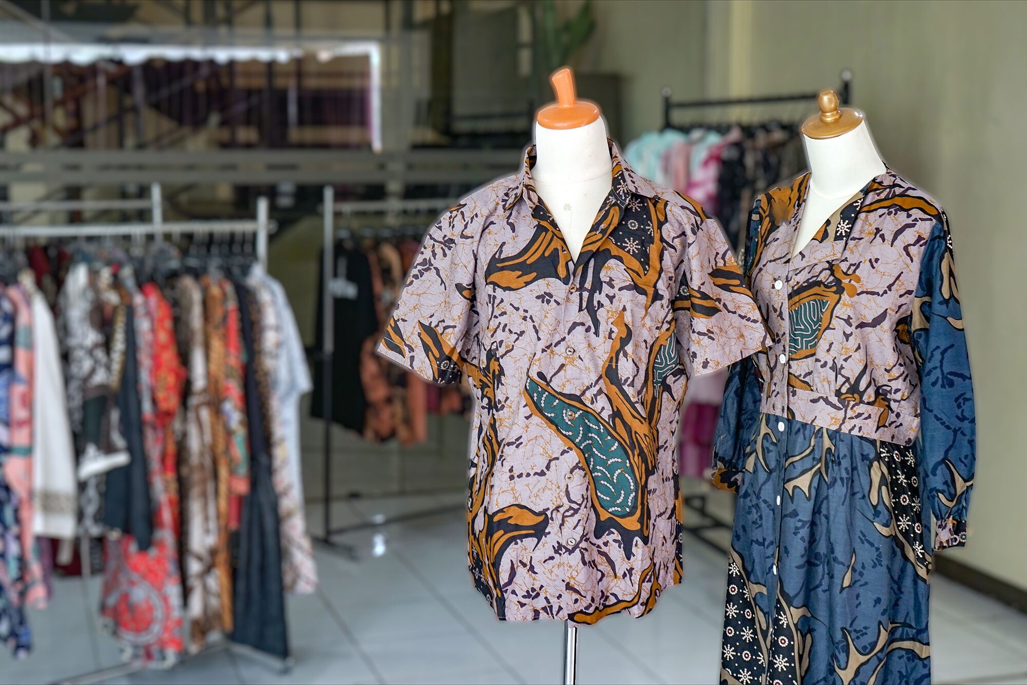 Two mannequins displaying patterned shirts in a retail setting with a rack of various clothing items in the background.