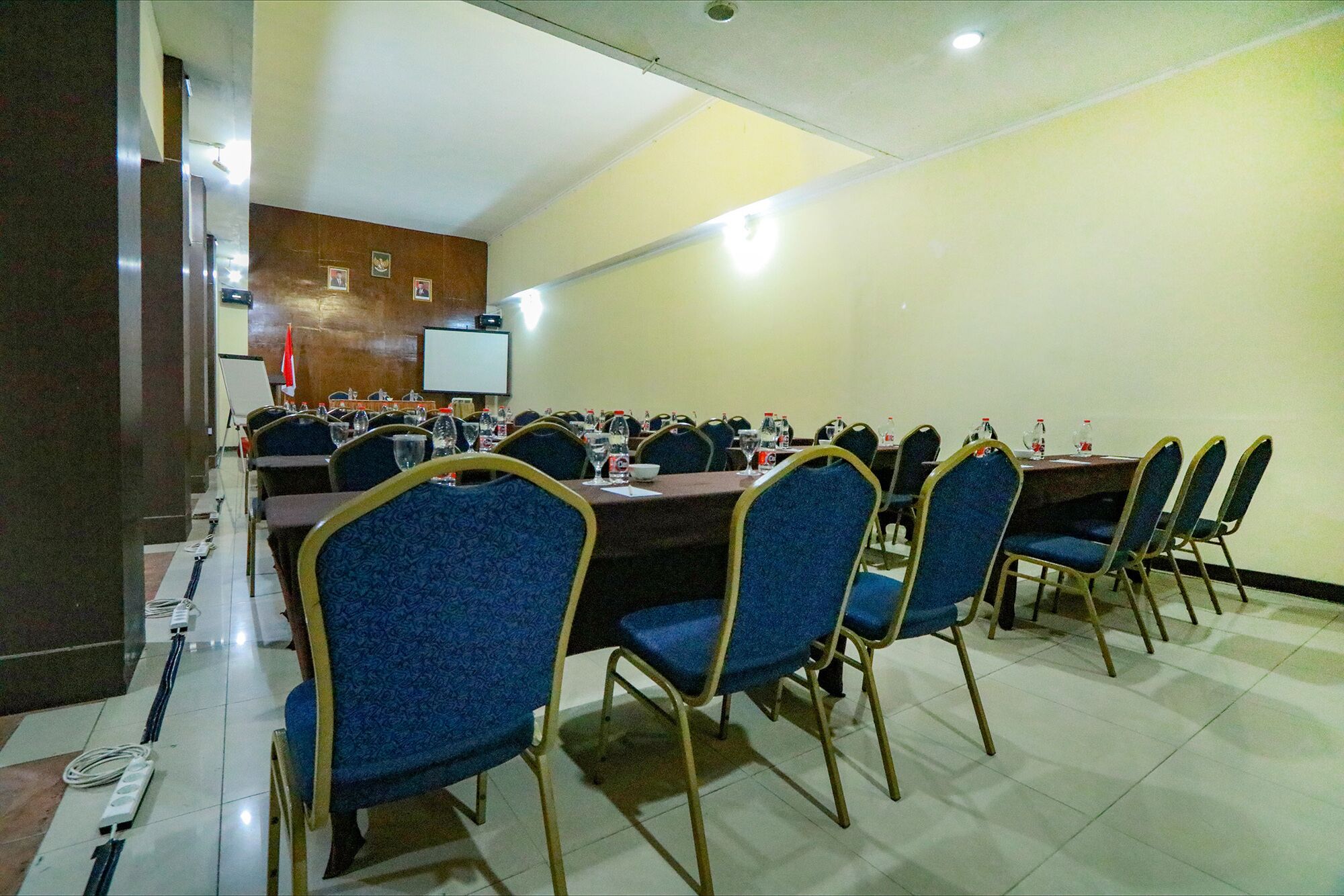 Un salón de conferencias con varias mesas y sillas, equipado con botellas de agua y una pantalla de proyector.