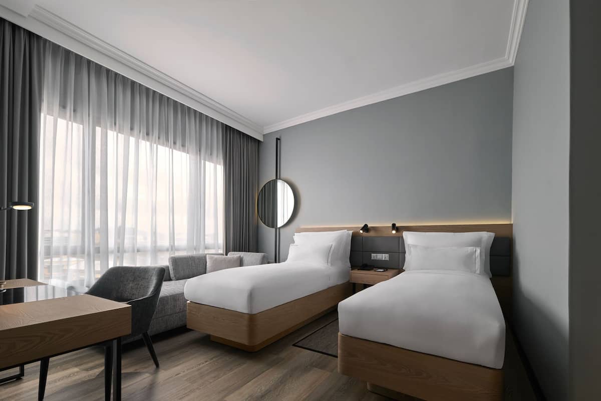 Una habitación de hotel moderna con dos camas, un escritorio, una silla y un ventanal con cortinas.