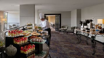 A hotel dining area with a chef preparing food, a buffet with various dishes, and a seating area with tables and chairs.