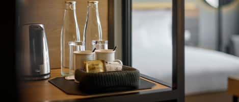 A neatly arranged hotel room with a wooden nightstand, a kettle, two glass bottles, a cup, and a small basket containing a tea bag.
