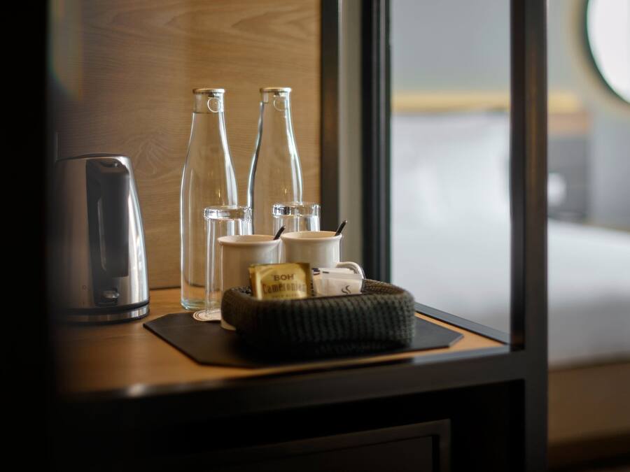 Habitación de hotel ordenada con una mesita de noche de madera, una tetera, dos botellas de vidrio, una taza y una pequeña cesta con una bolsita de té.