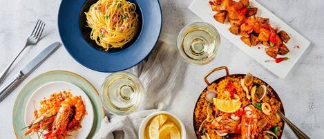 A dining table with a plate of pasta, a pan of seafood paella, and a plate of shrimp.