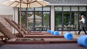 Een zwembadgedeelte met ligbedden en parasols, een eethoek met tafels en stoelen, en een persoon die langskomt.