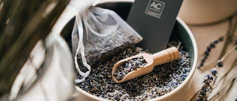 A bowl of dried lavender with a wooden scoop and a small sachet, placed on a textured surface with scattered lavender branches.