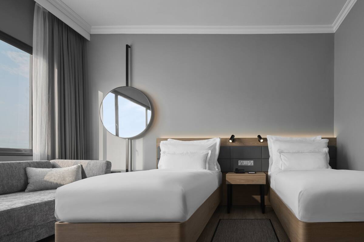 Een moderne hotelkamer met twee enkele bedden, een ronde spiegel, een nachtkastje met lamp, een bank en een groot raam met uitzicht.