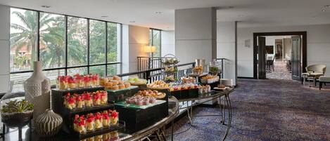 A spacious room with a long buffet table filled with various food items, large windows with a view of palm trees, and a carpeted floor.