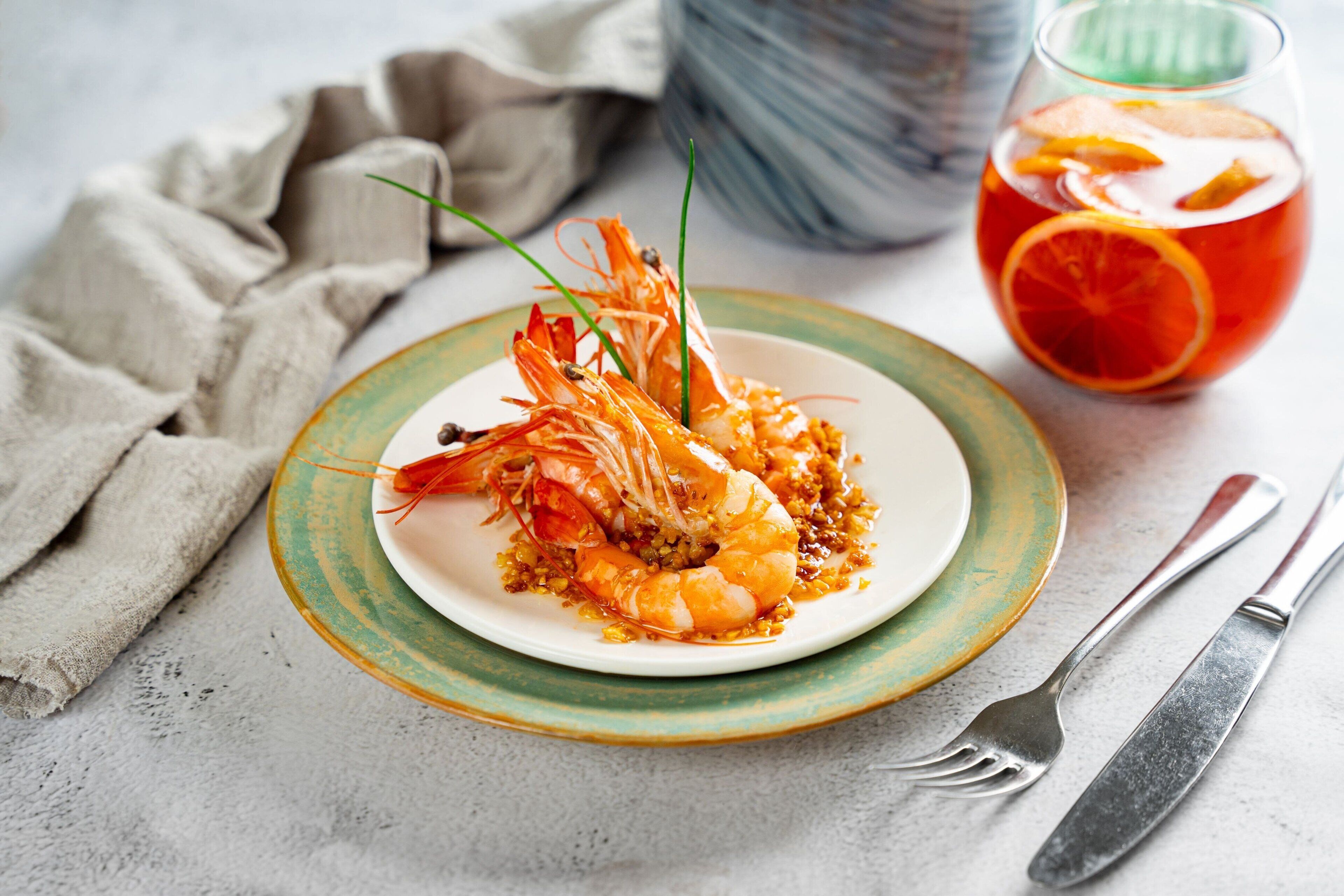 A plate of cooked shrimp with a garnish, a glass of iced drink with orange slices, and a fork and knife on a table.