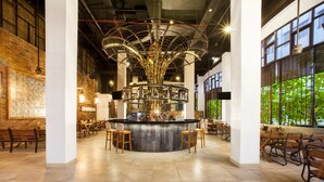 A modern bar area with a central chandelier, seating around the bar, and large windows with greenery views. - Sun Island Hotel & Spa Legian (Kuta)