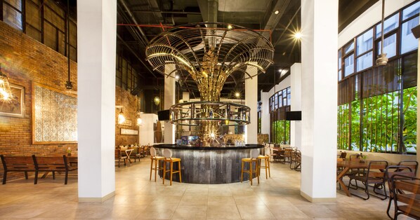A modern bar area with a central chandelier, seating around the bar, and large windows with greenery views.