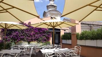 An outdoor patio with white metal chairs and tables, covered by large beige umbrellas, and a view of a building with a domed roof and a bell tower.