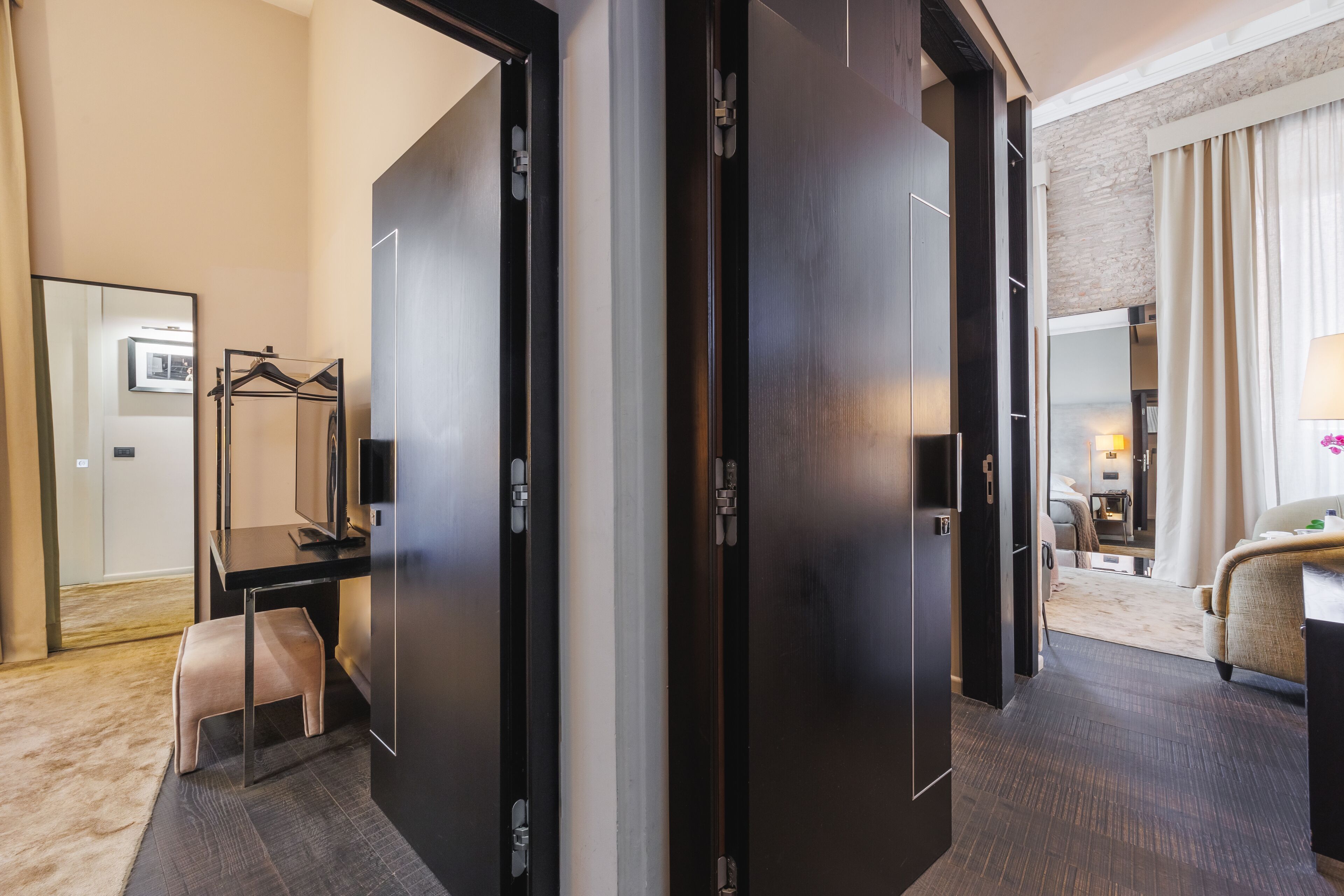 A modern hotel room with a dark wood wardrobe, a desk with a chair, a small bench, and a view into the living area.