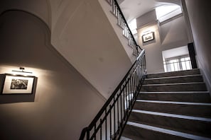 A grand staircase with marble steps and black balusters, leading to a landing with a framed picture and a light fixture.