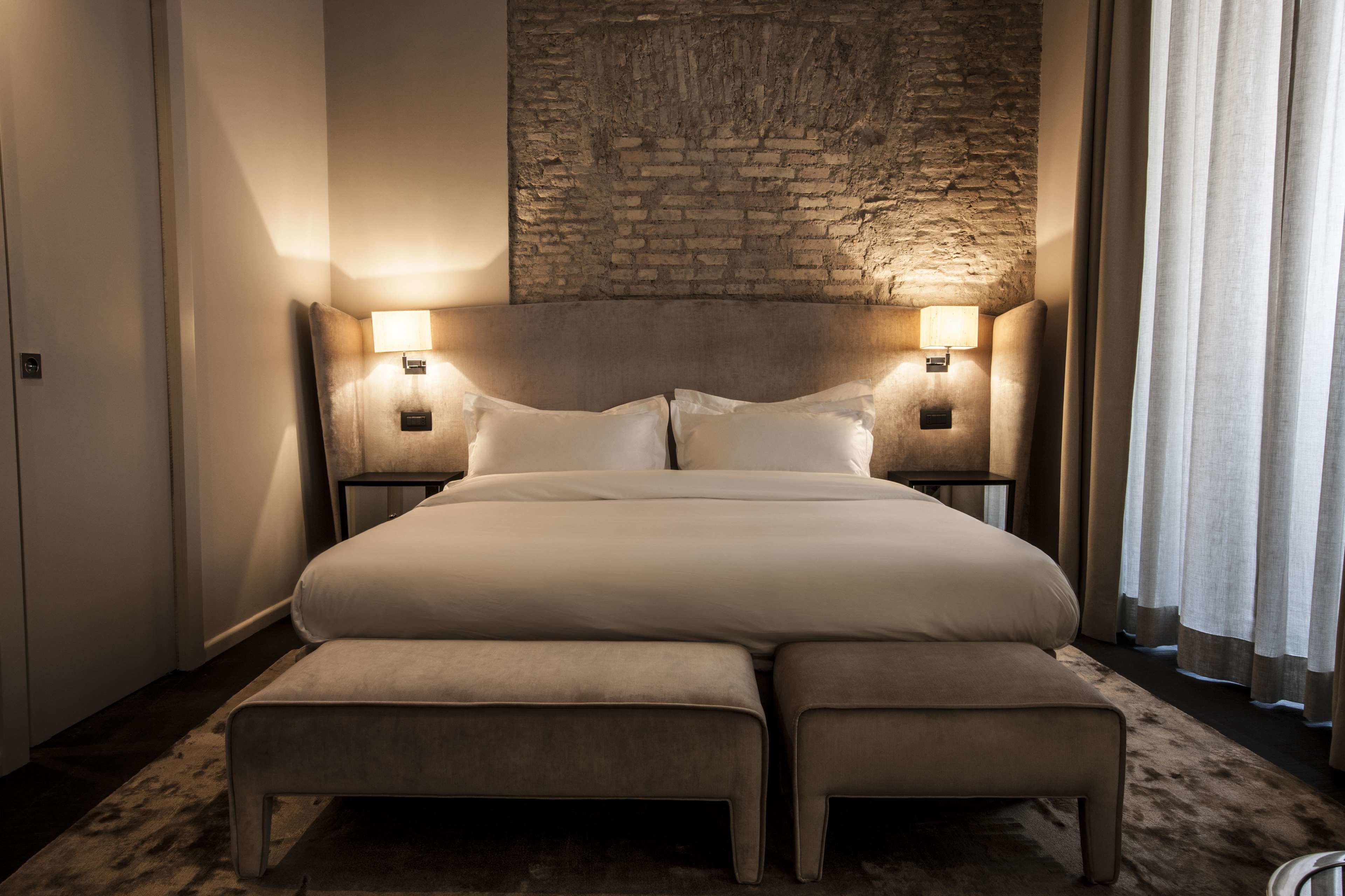 Una cama grande con dos mesitas de noche, un banco y una pared de piedra al fondo.