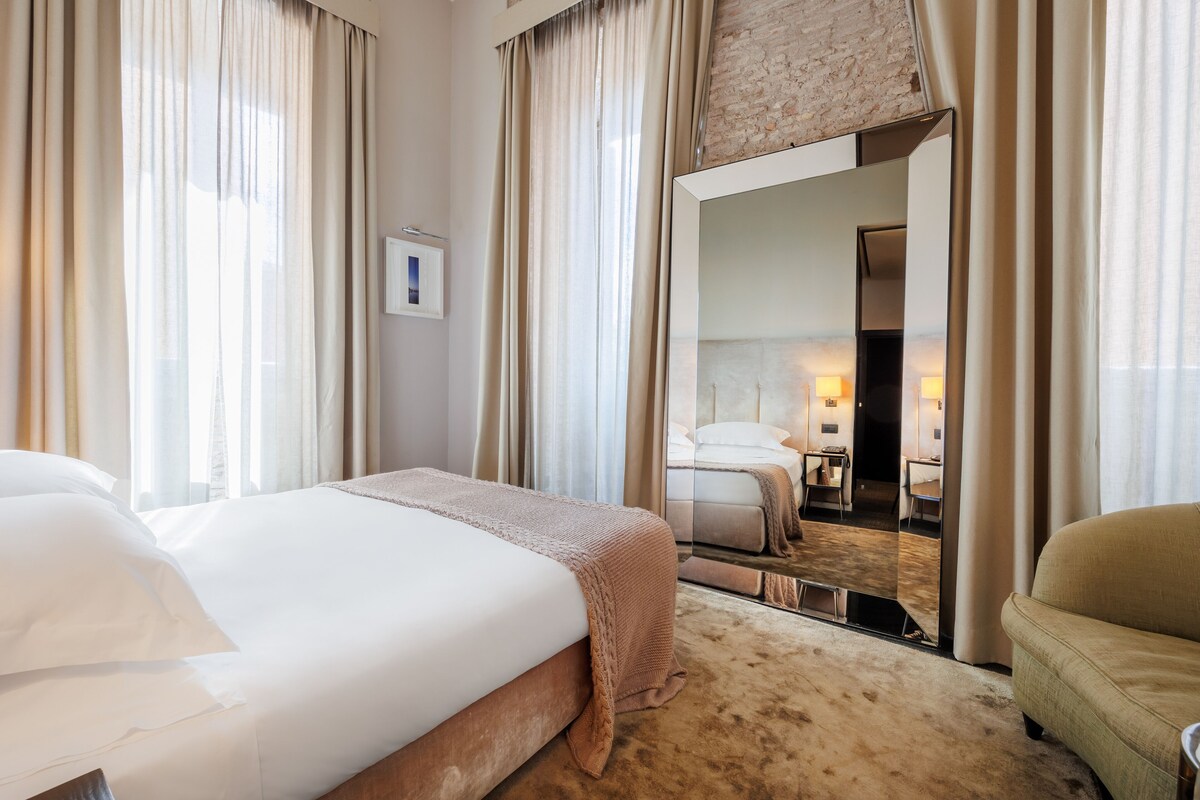 A hotel room with a large bed, a sitting area with a sofa, a mirror, and a view of a bathroom with a shower and toilet.