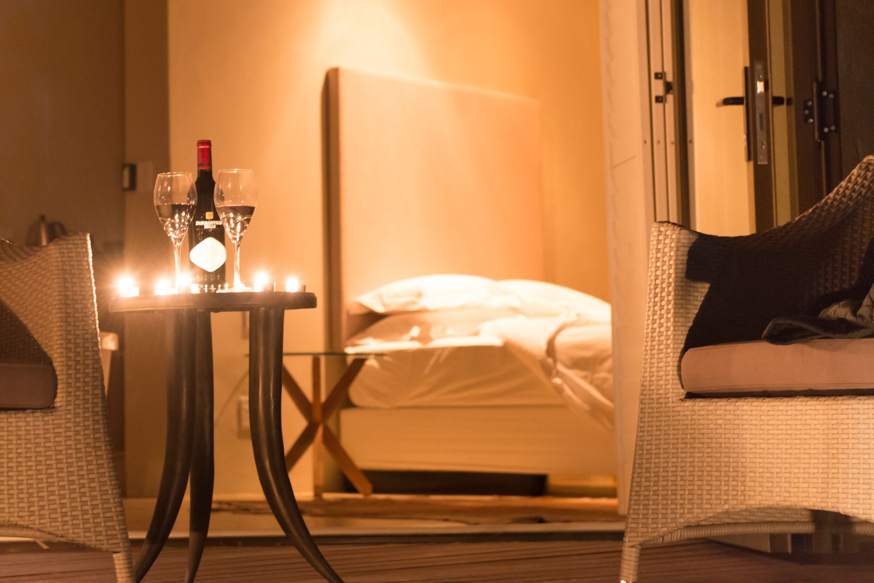 A hotel room with a bed, a small table with two glasses of wine, and a chair.
