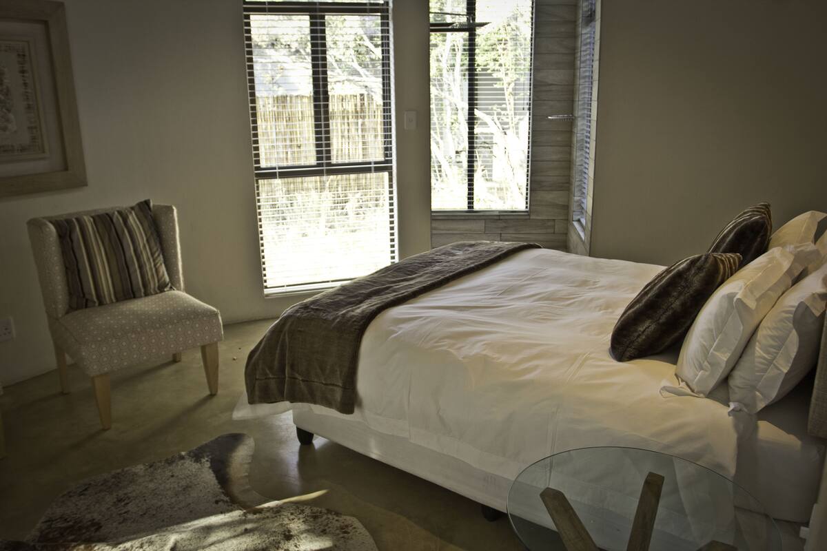 A bedroom with a large bed, a chair, and two windows with blinds.