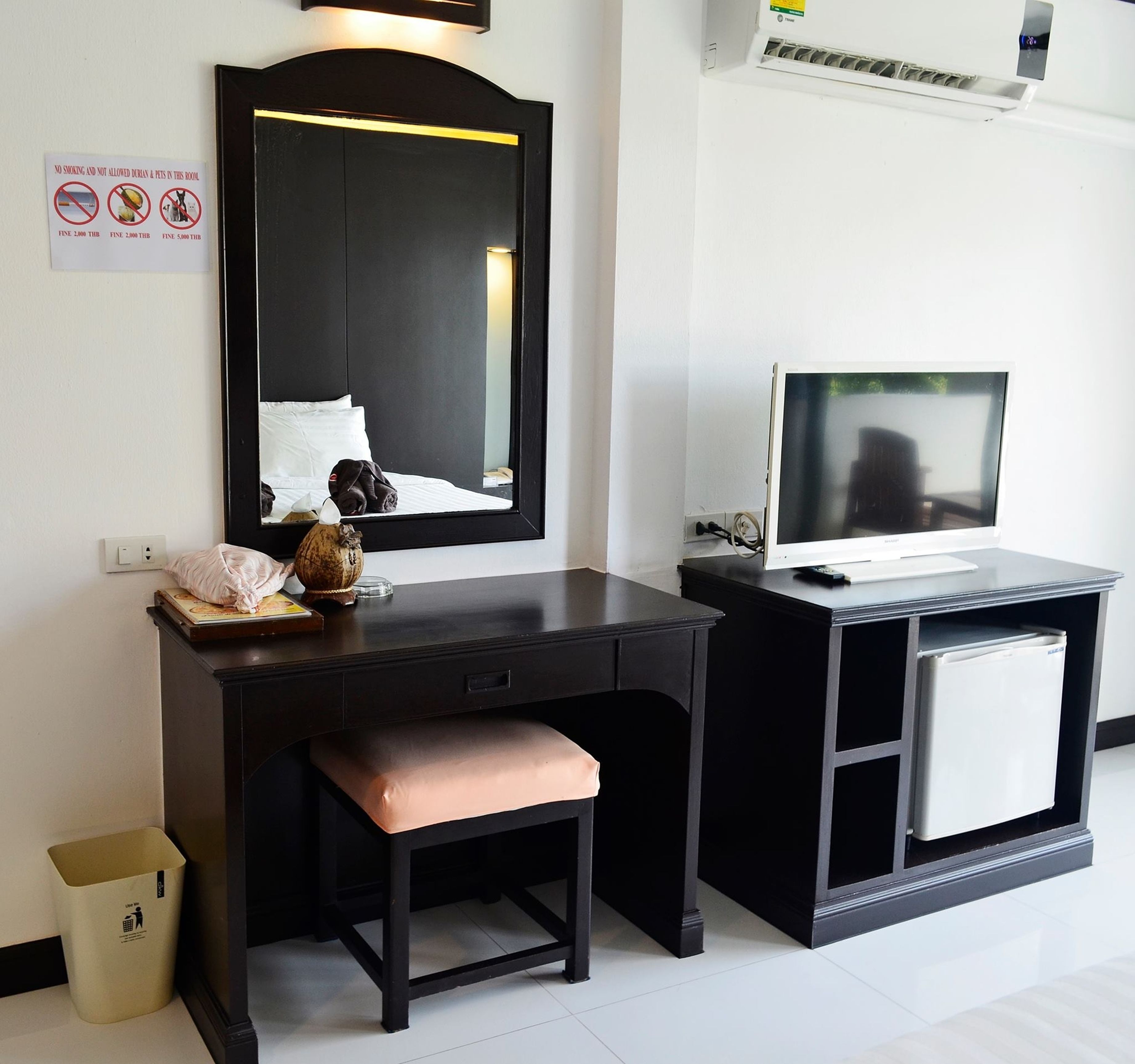 Quarto de hotel com escrivaninha preta, um banquinho, um espelho, televisão e lixeira.