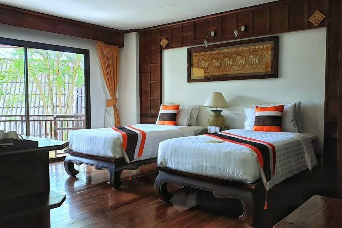 A hotel room with two beds, a desk, and a large window with a view of trees.