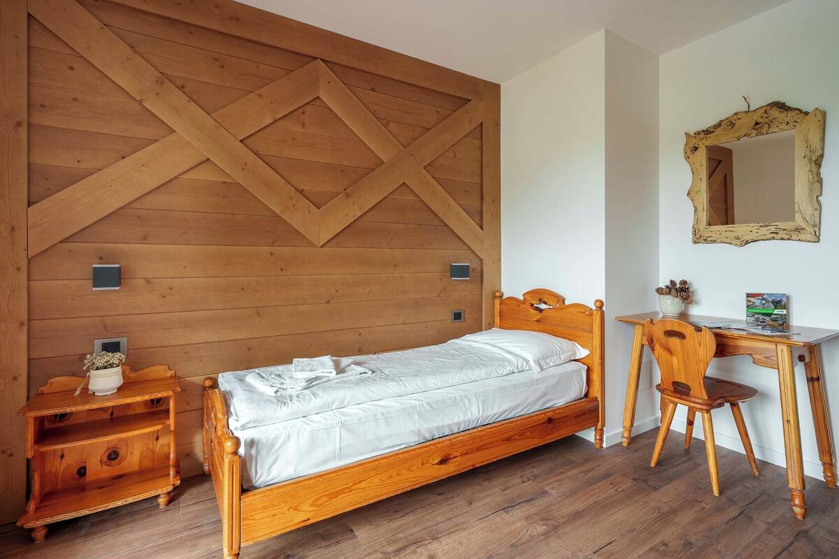 A wooden bed with a headboard, a wooden nightstand, a wooden desk with a chair, and a mirror on the wall.