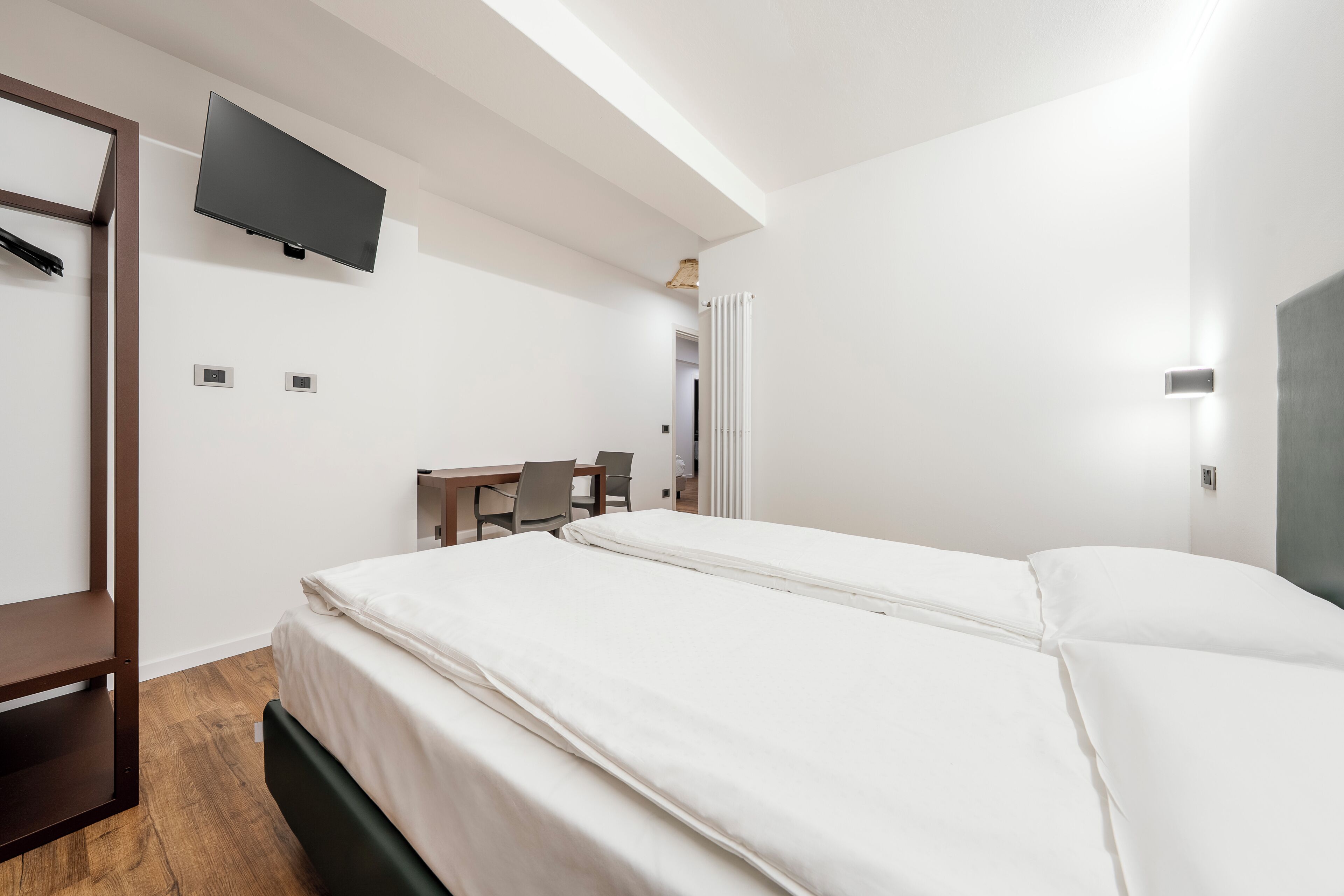 A hotel room with a bed, a desk, a chair, a TV mounted on the wall, and a radiator.
