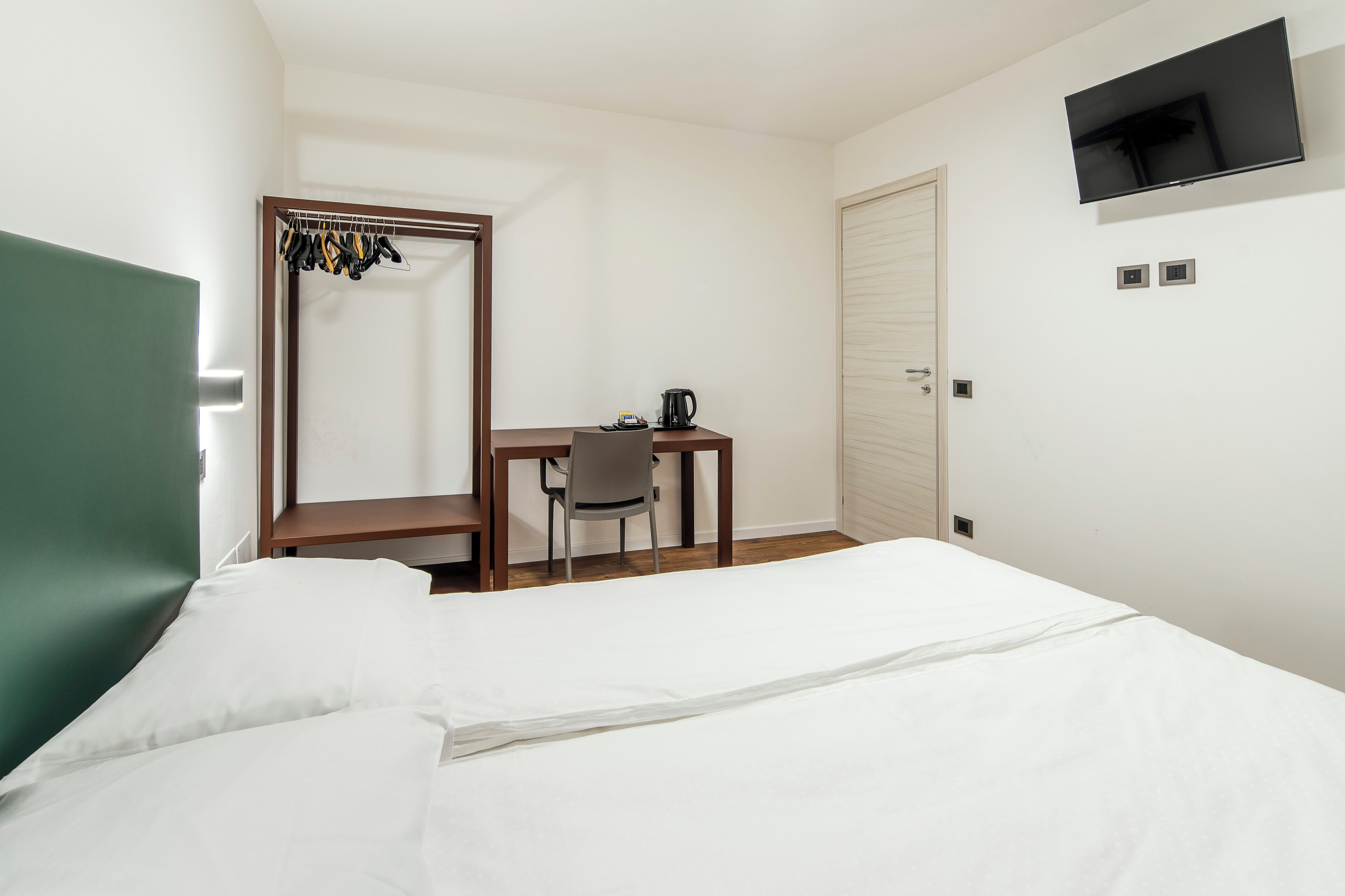 A hotel room with a bed, a desk with a chair, a coat rack, a television, and a door.