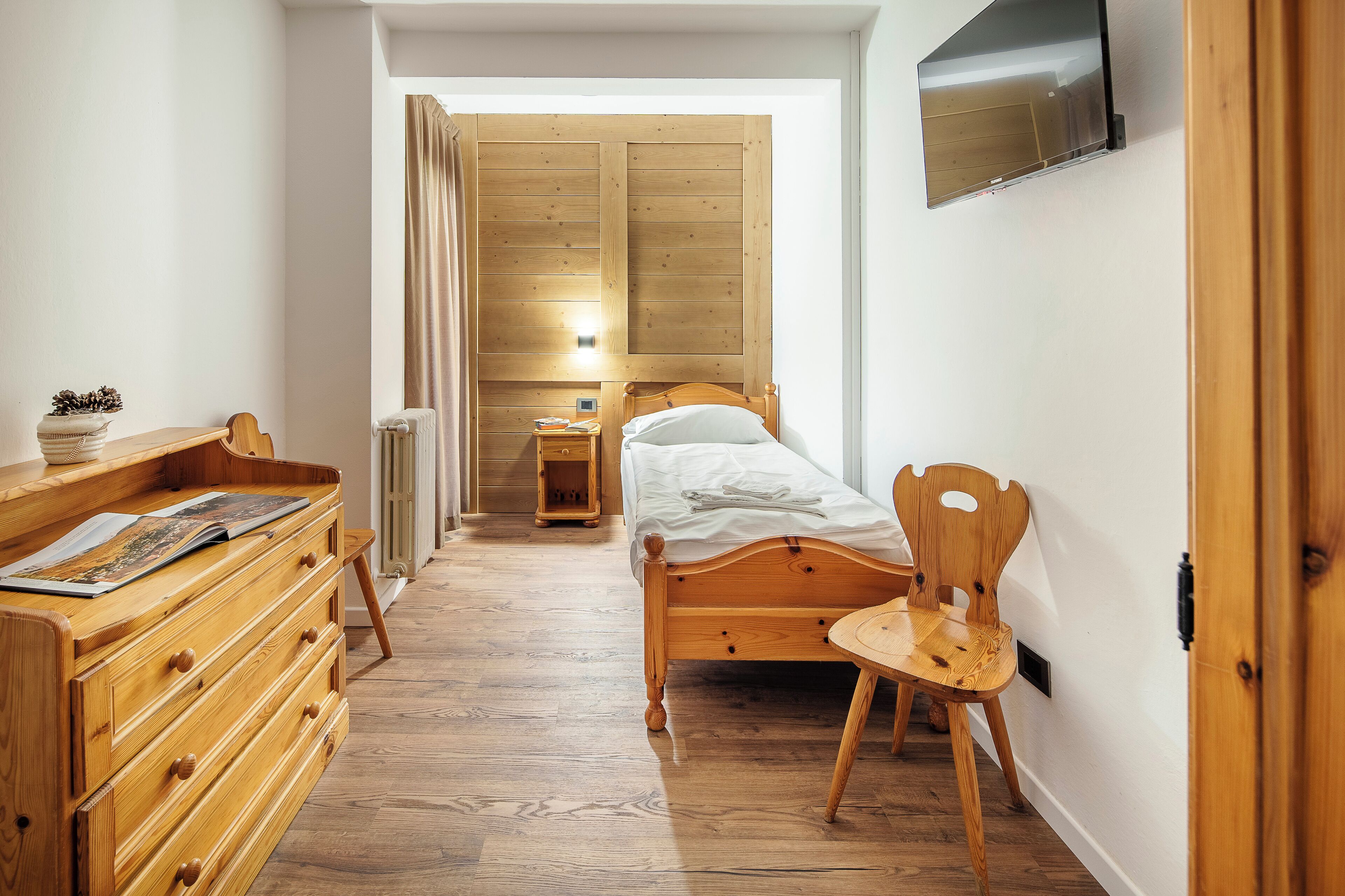 Une chambre à coucher avec un lit en bois, une commode en bois, une chaise, une table de chevet et un téléviseur fixé au mur.