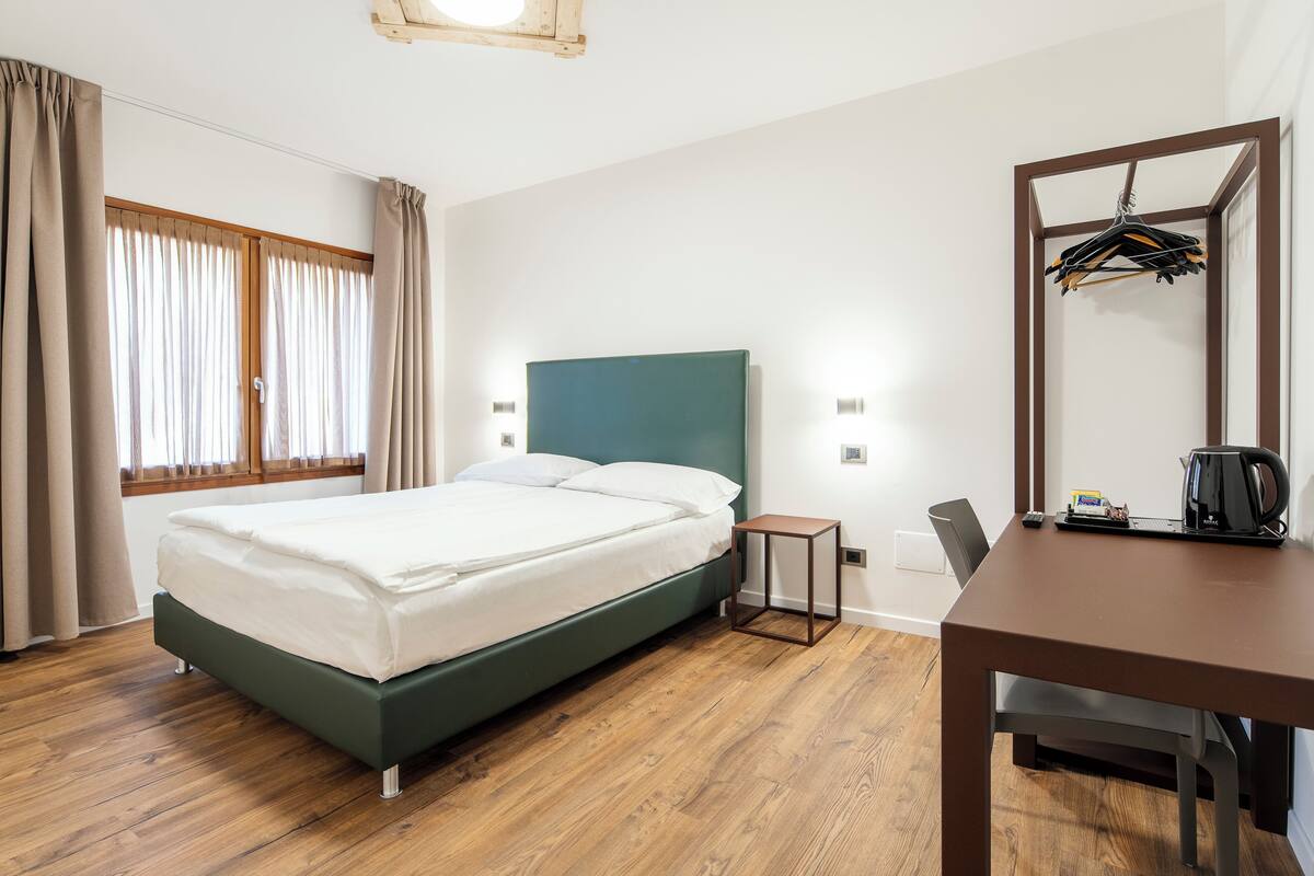 A bedroom with a bed, wooden floor, a desk, a chair, a mirror, and a window with curtains.