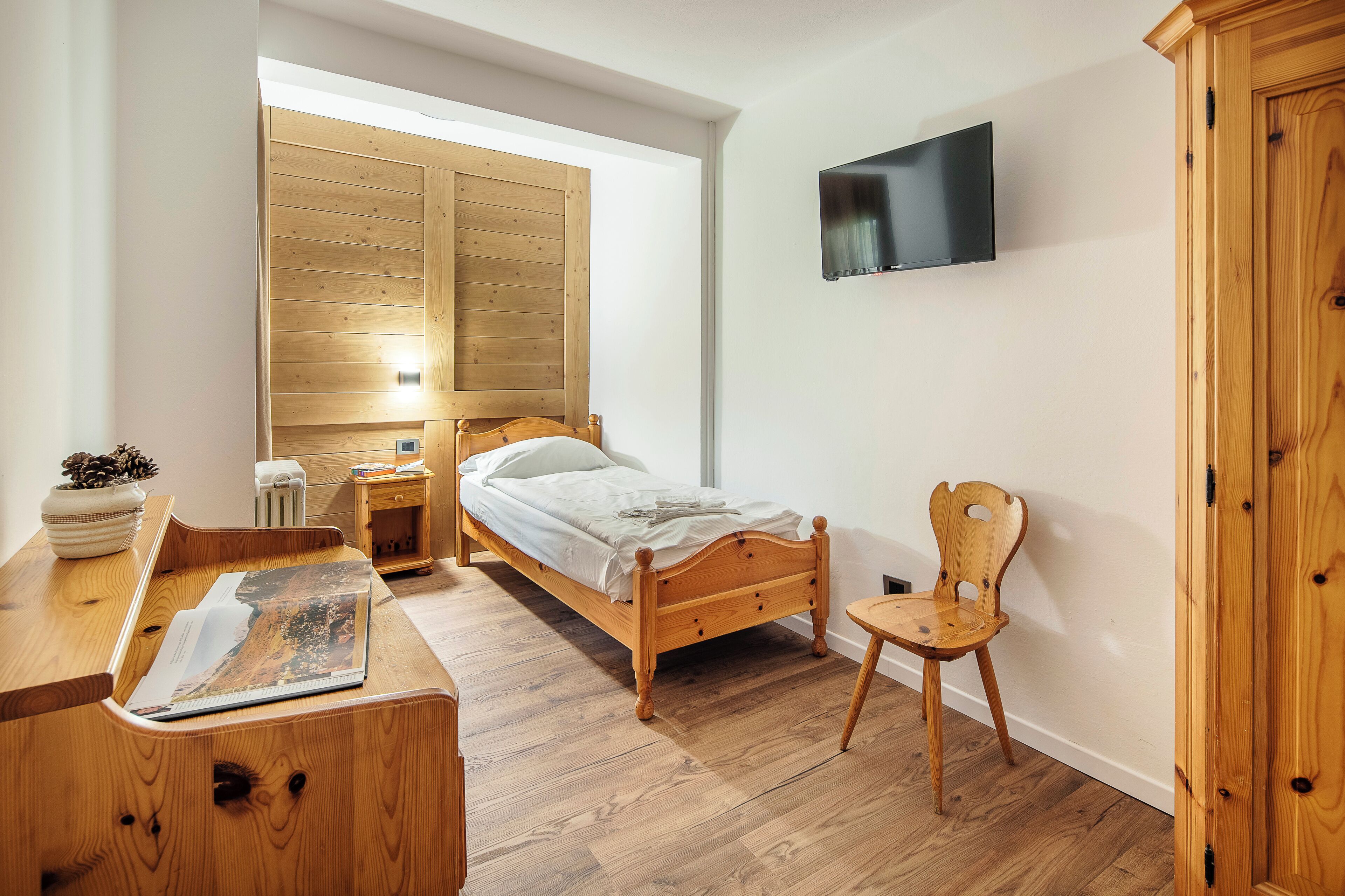 Ein Schlafzimmer mit einem Holzbett, einem an der Wand montierten Fernseher, einem Holzstuhl und einem Schreibtisch mit einer Zeitschrift.