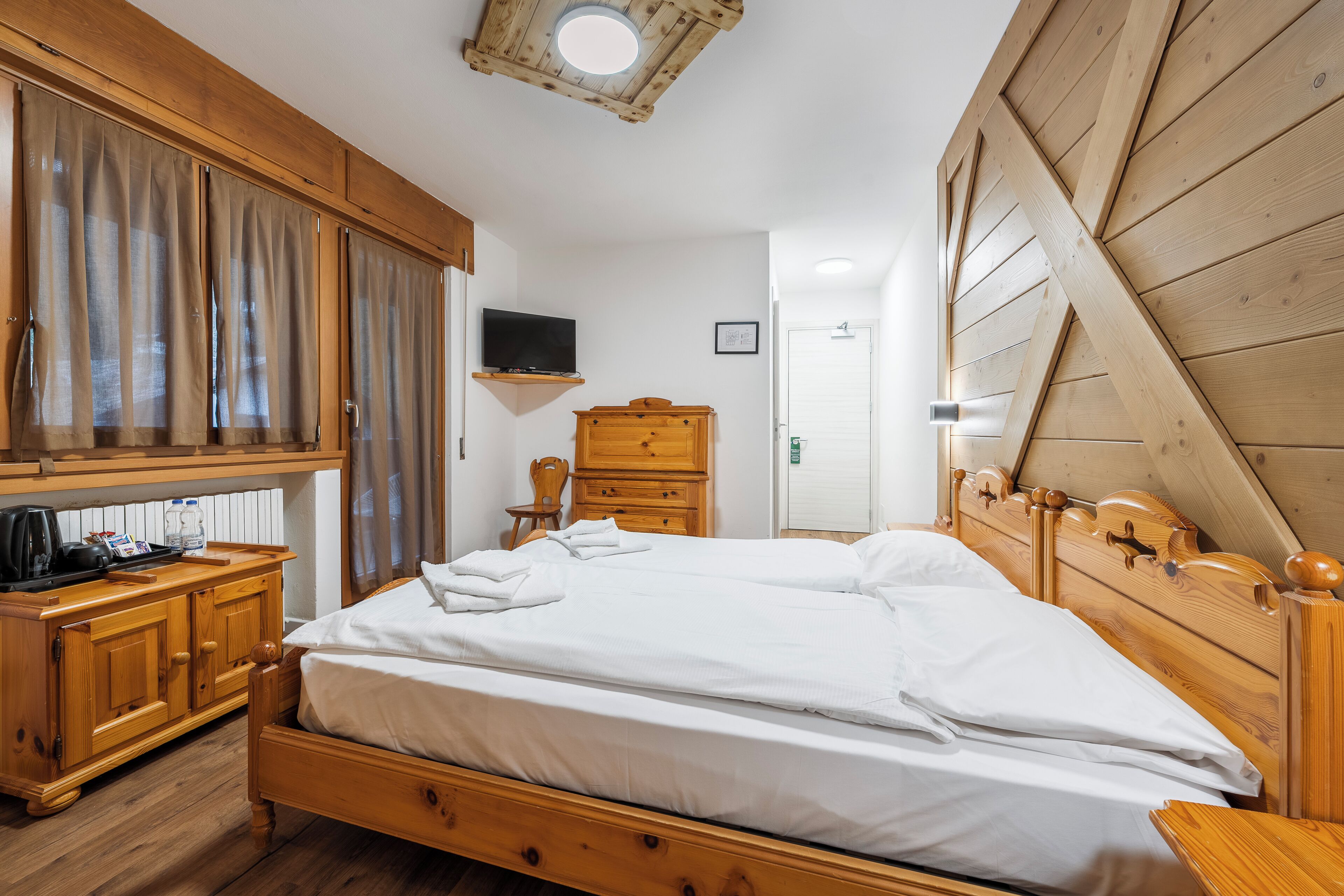 A wooden-framed bedroom with a bed, a wooden dresser, a television on a shelf, and a door.