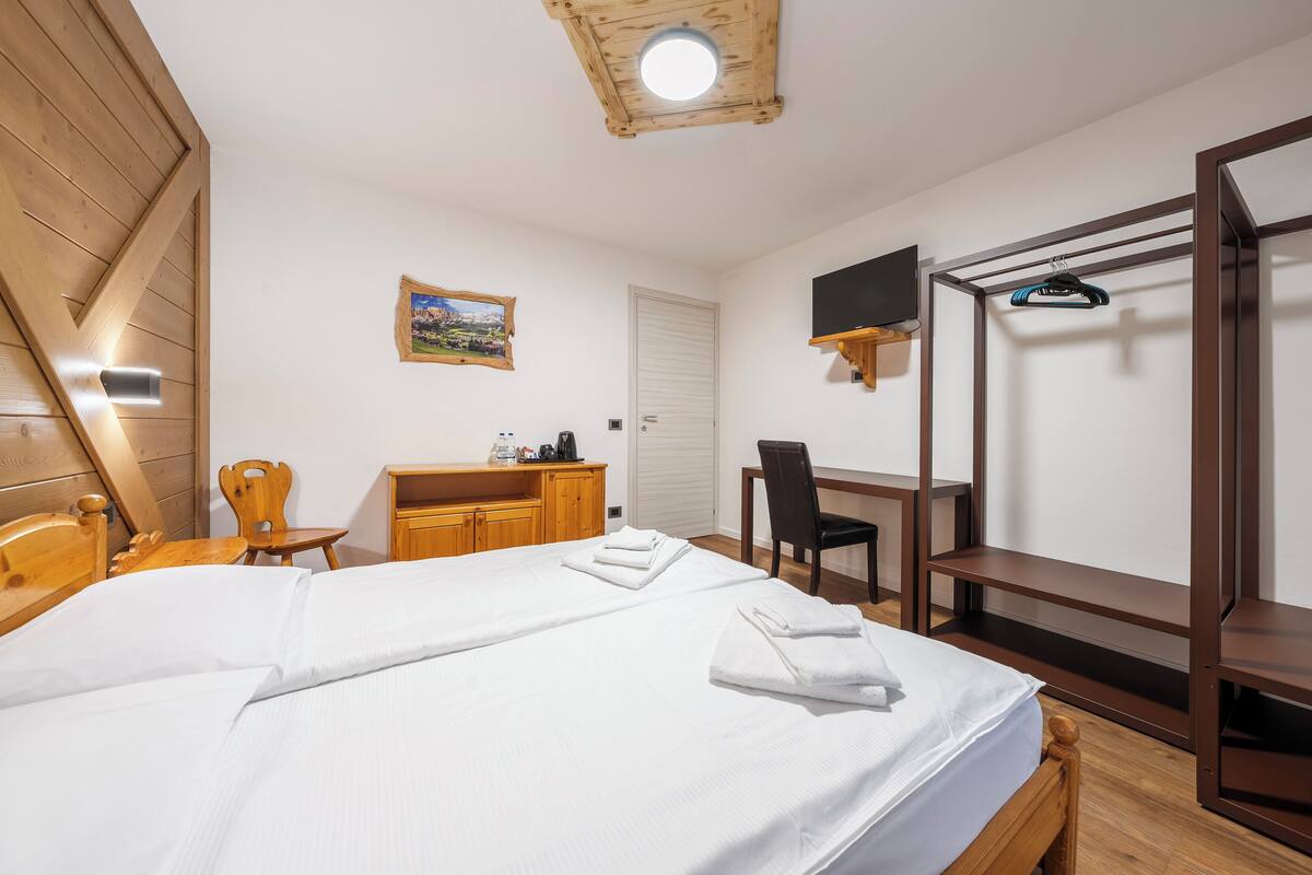 A bedroom with a bed, wooden headboard, a desk with a chair, a TV mounted on the wall, and a wooden wardrobe.